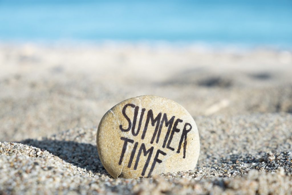 Announces our summer hours with a closeup of a round stone with the text summer time written in it, on the sand of a beach, with the sea in the background